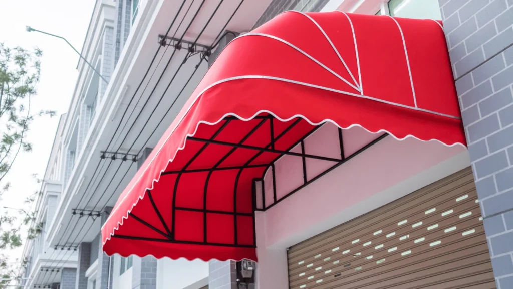 A red fabric awning with white trim and black metal supports extends over the entrance of a modern building, providing shade above a closed roller shutter.