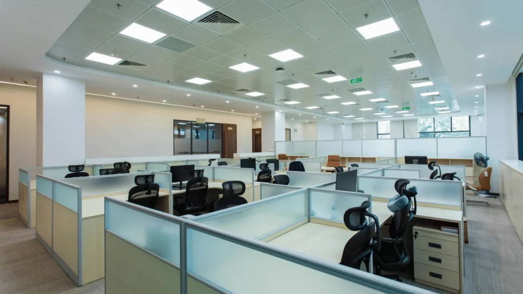 Bright, spacious modern open-plan office with rows of partitioned workstations, ergonomic office chairs, desks, and overhead fluorescent lighting.